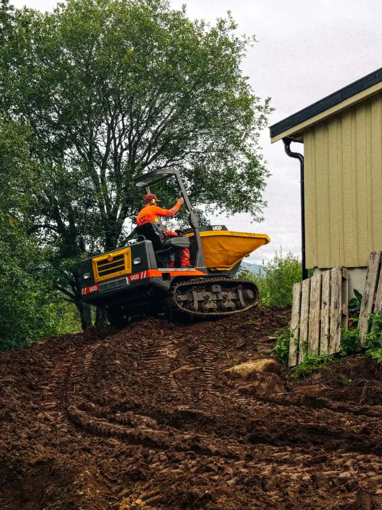 Beltedumper kjører jordmasser opp en bratt skråning ved huset