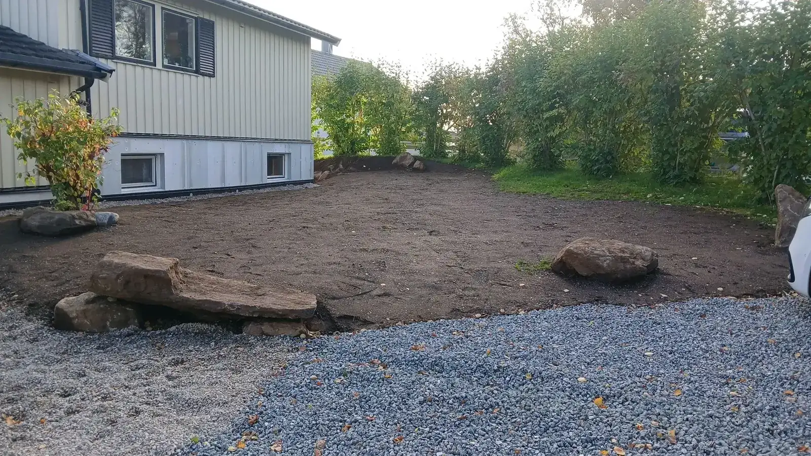 Planert jordflate avgrenset av grus og store steinblokker, klar for plen og beplanting